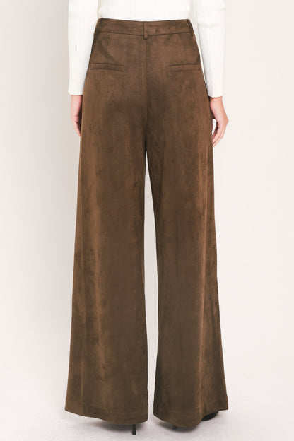 A person shown from the back wearing high-waisted, wide-leg brown pants with a suede-like finish, paired with a white long-sleeve top against a plain light background.
