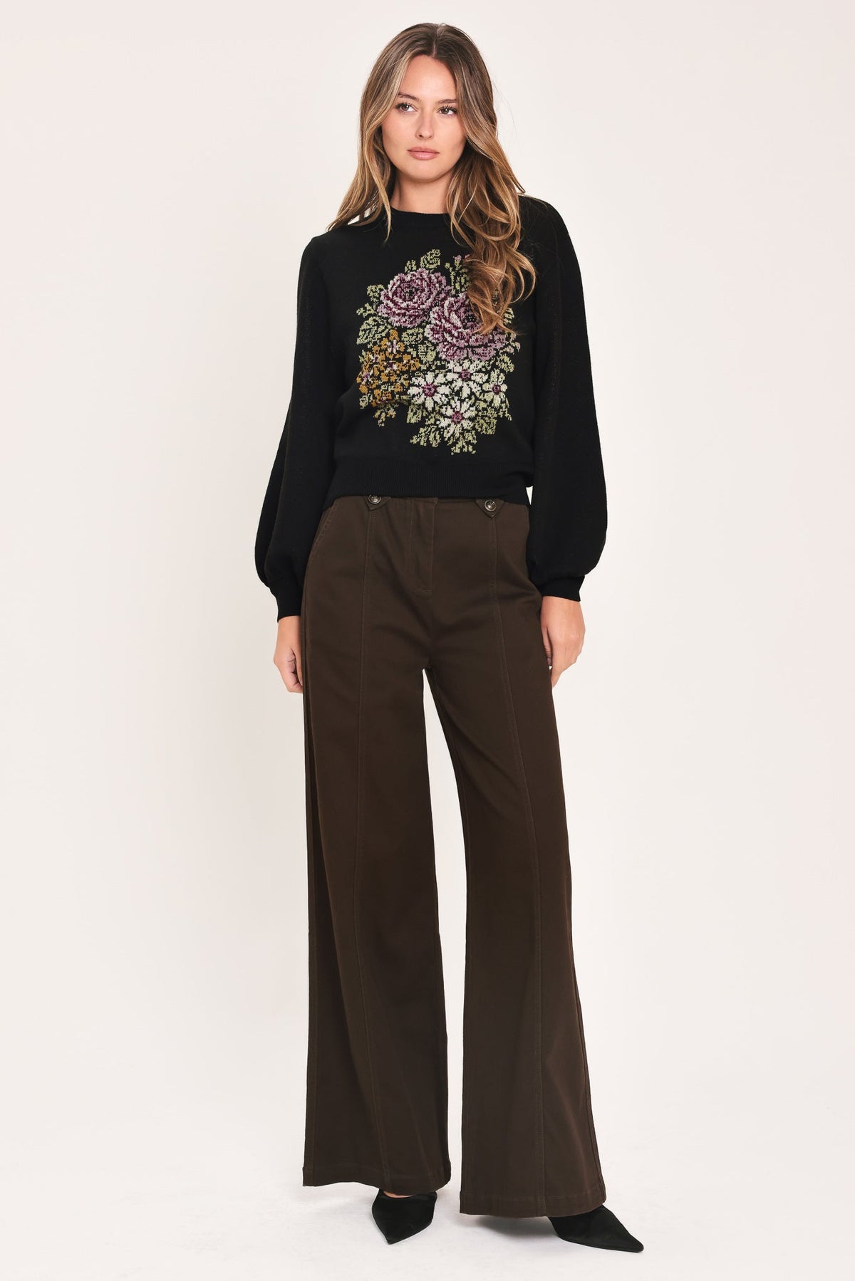 A woman modeling a black long-sleeve knit sweater with a colorful floral embroidery on the chest, paired with dark brown wide-leg trousers, against a plain light background.