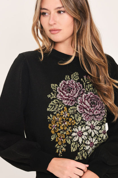 A woman wearing a black crewneck sweater with a large, colorful floral embroidery design on the front, featuring pink, purple, yellow, and white flowers with green leaves.