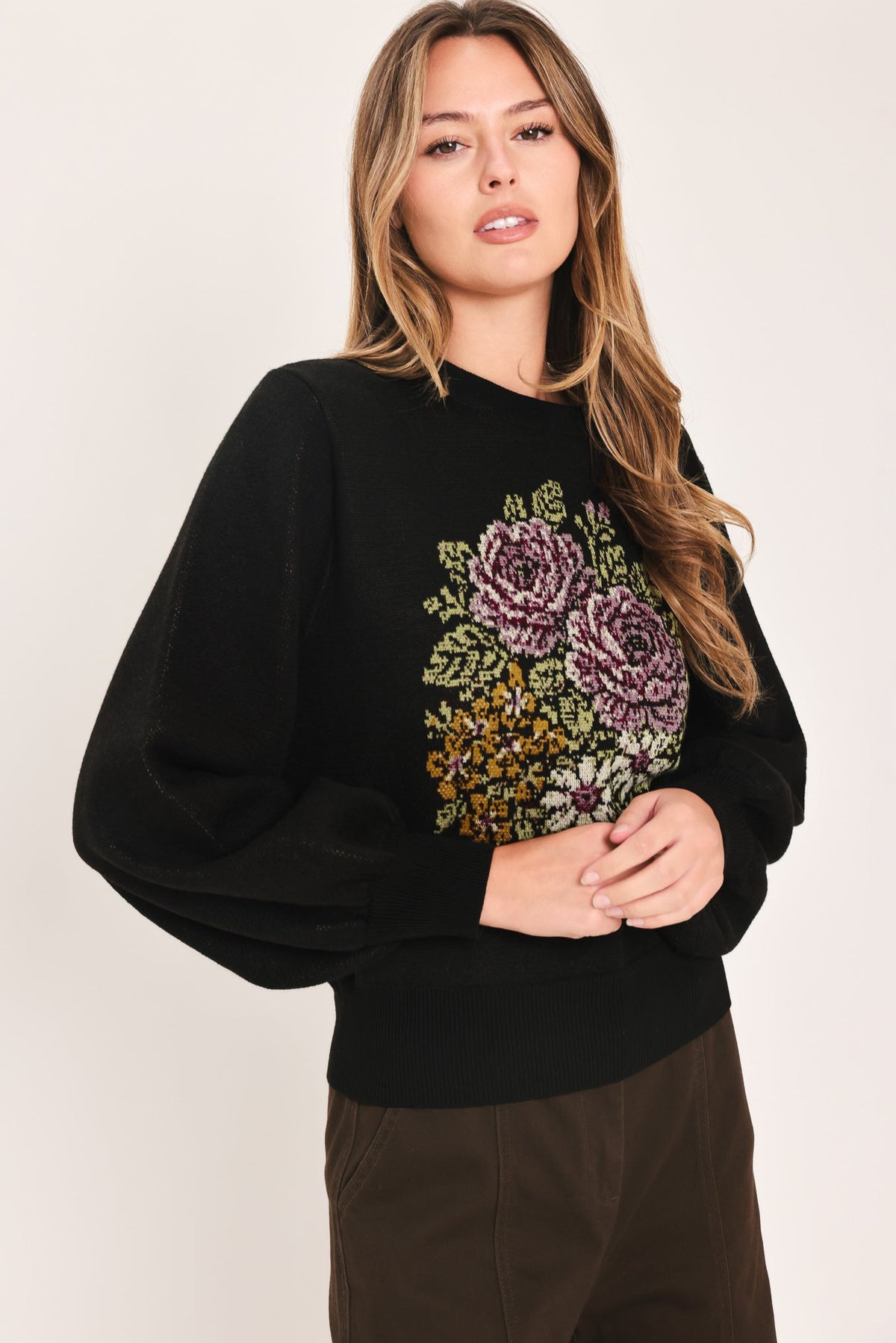 A woman wearing a black crewneck sweatshirt with a large, colorful floral embroidery design on the front, paired with brown pants, against a plain light background.