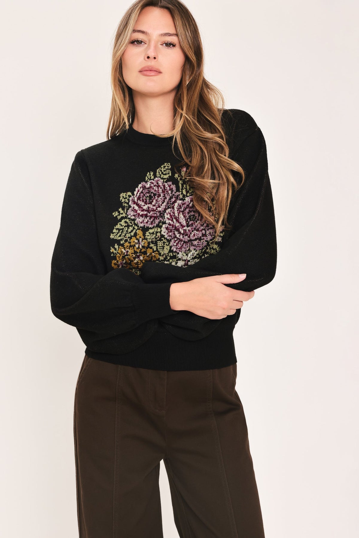 A woman modeling a black crewneck sweatshirt featuring a large embroidered floral design in purple, green, and gold on the front, paired with dark brown trousers, against a plain light background.