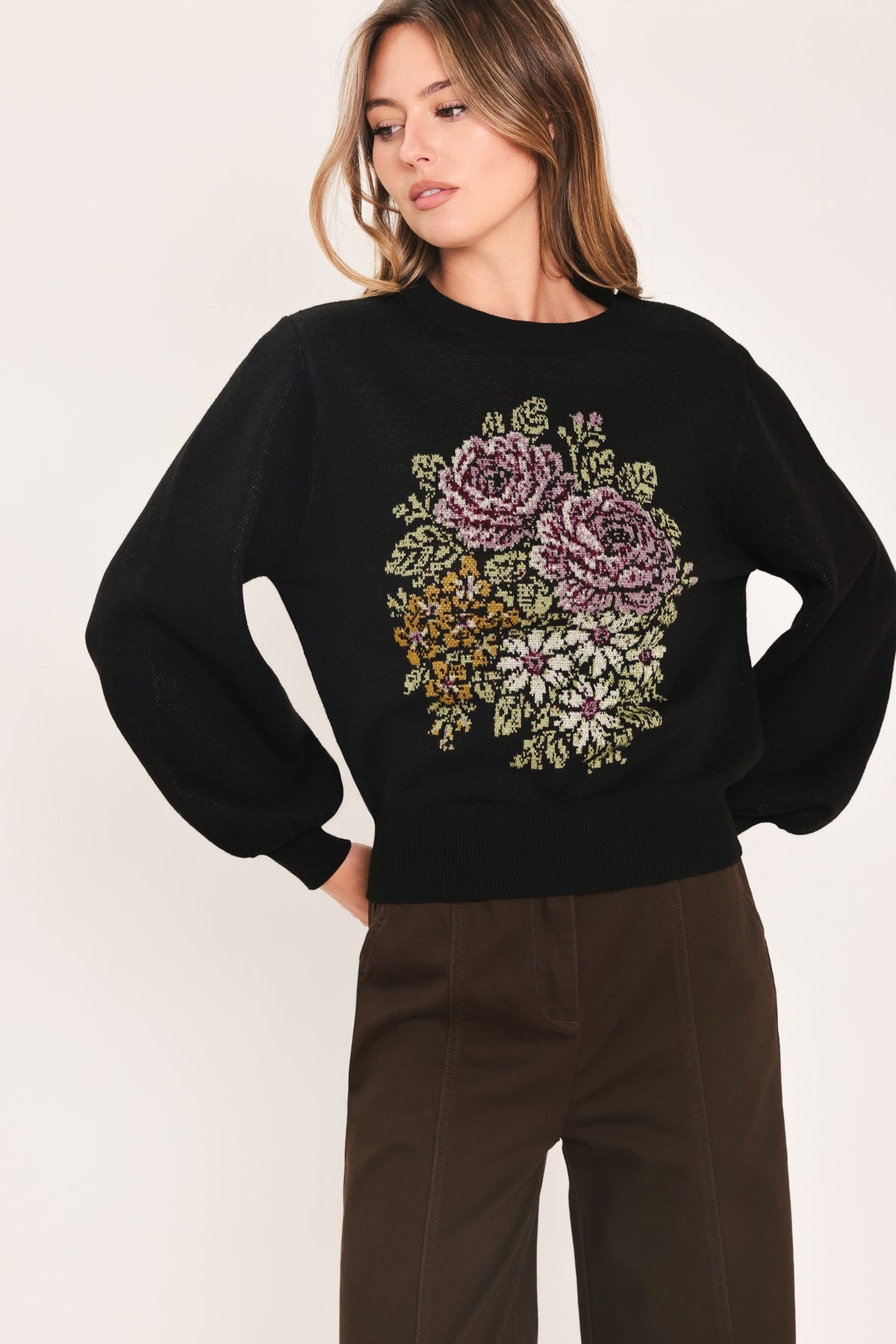 A woman wearing a black crewneck sweatshirt with a large, colorful floral embroidery design on the front, paired with dark brown trousers, against a plain light background.