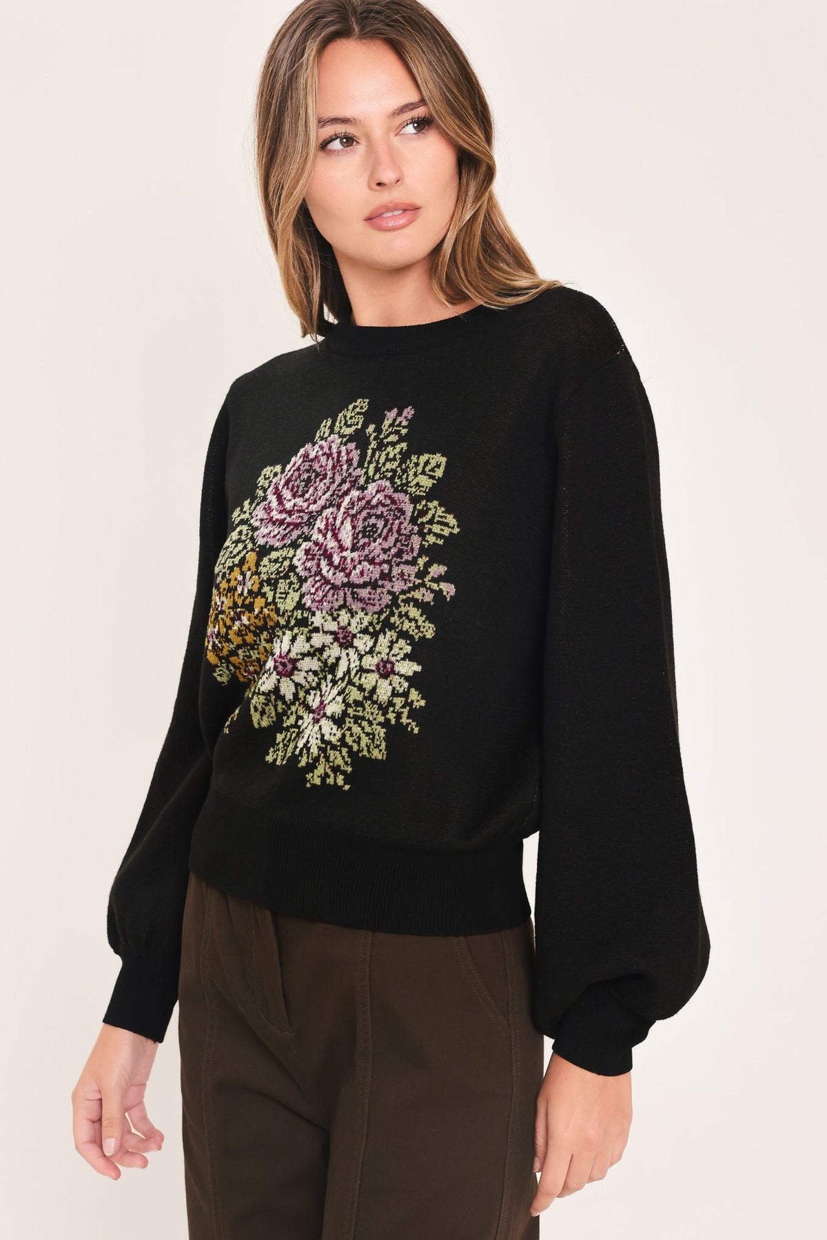 A woman wearing a black long-sleeve knit sweater with a large embroidered floral design in purple, green, and gold on the front, paired with dark brown pants, against a plain light background.