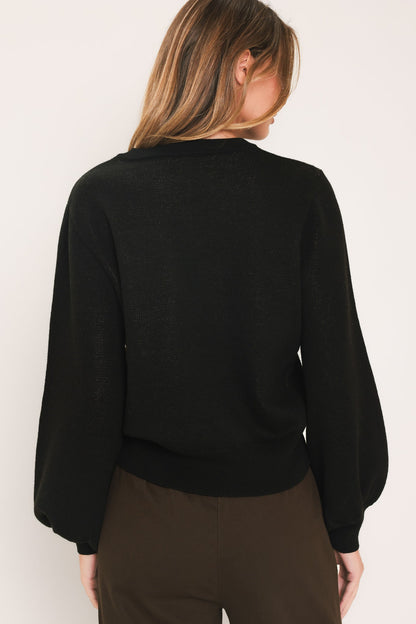 A woman shown from the back wearing a black long-sleeve knit sweater and dark brown pants against a plain off-white background.