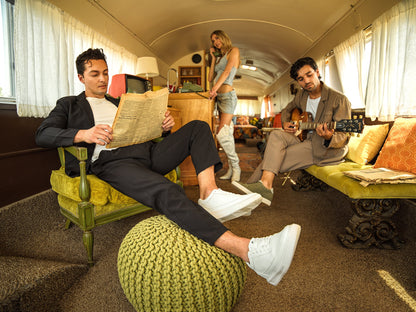 People inside a vintage-style bus living room with one person reading a newspaper, another playing guitar, and a third person standing.