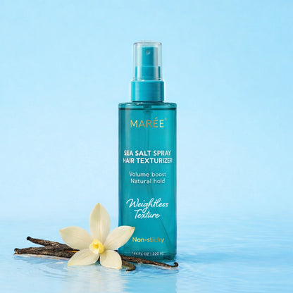A bottle of MARÉE Sea Salt Spray Hair Texturizer stands on water with a white flower and vanilla beans beside it against a light blue background.
