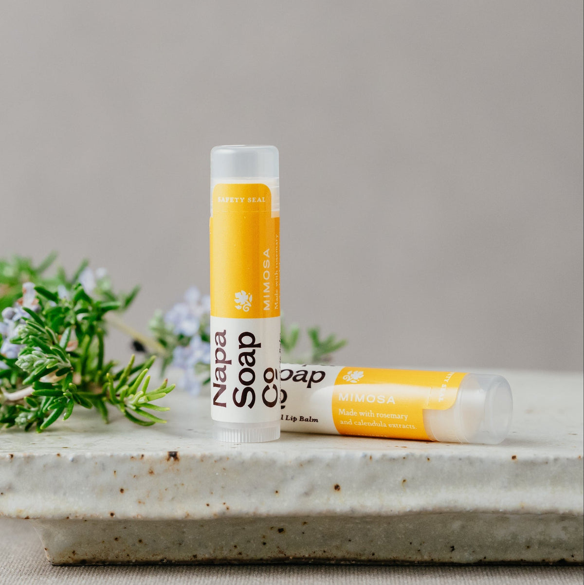 Two lip balms with visible branding on a stone surface with greenery in the background