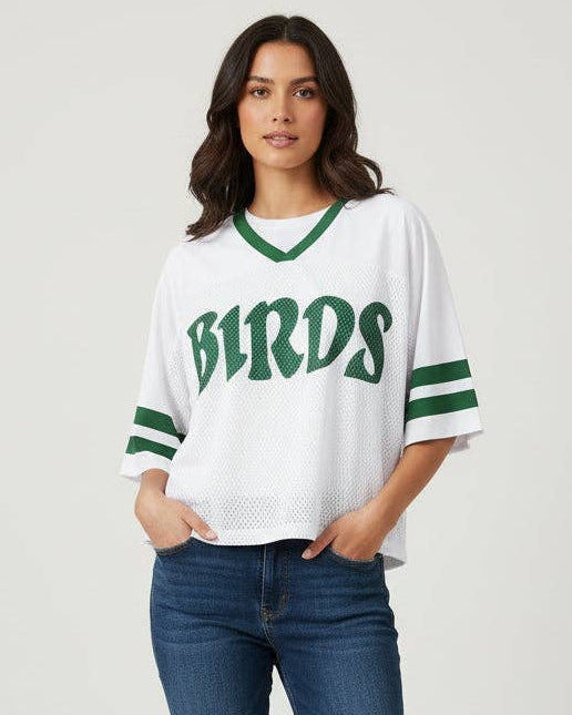 A woman modeling a white mesh V-neck jersey top with green trim, green 'BIRDS' lettering on the chest, and green striped sleeve accents, paired with blue jeans against a light gray studio background.