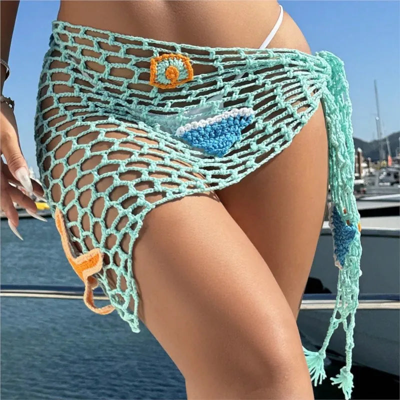 Person wearing a green crochet cover-up with decorative elements by a body of water.