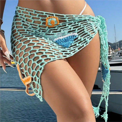 Person wearing a green crochet cover-up with decorative elements by a body of water.