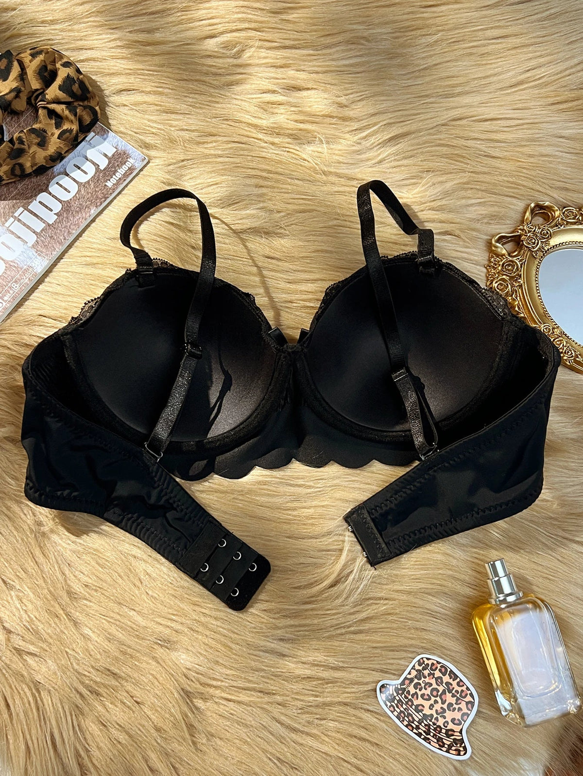 A black lace and mesh underwire bra with wide shoulder straps and a scalloped underband, laid flat on a beige faux fur surface with decorative props including a leopard-print hair scrunchie, a magazine, a gold mirror, and a perfume bottle.