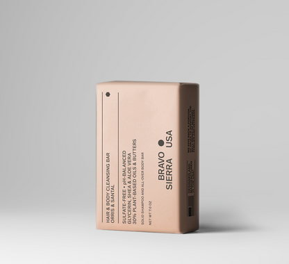 A single rectangular hair and body cleansing bar package standing upright on a clean light gray studio background. The package is a warm blush-beige color with black text reading BRAVO SIERRA USA and product details including coconut, shea, and argan oils.
