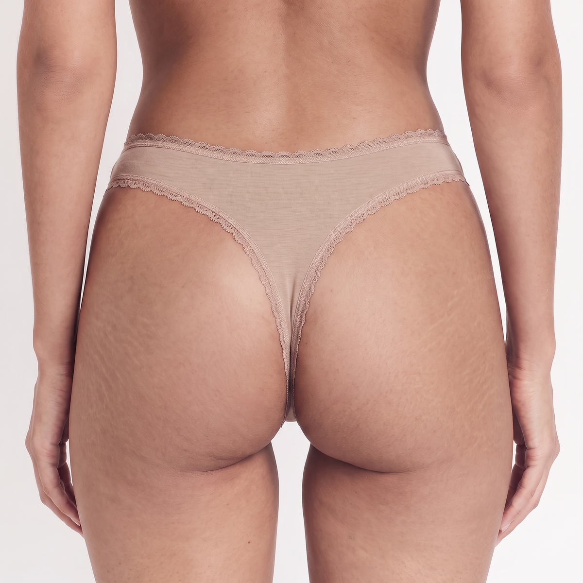 Rear view of a model wearing a beige thong with delicate lace trim on a light gray studio background.
