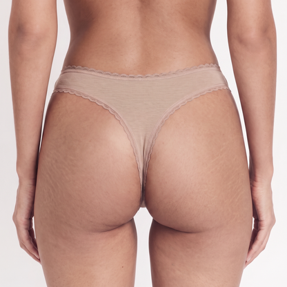 Rear view of a model wearing a beige thong with delicate lace trim on a light gray studio background.