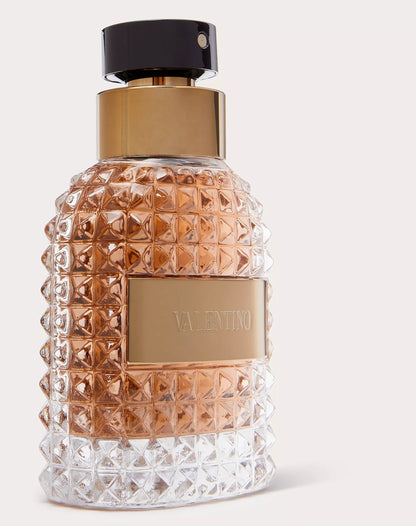 Valentino perfume bottle with a textured clear glass body, rose gold liquid, a gold-tone collar, and a black spray top on a light gray background.