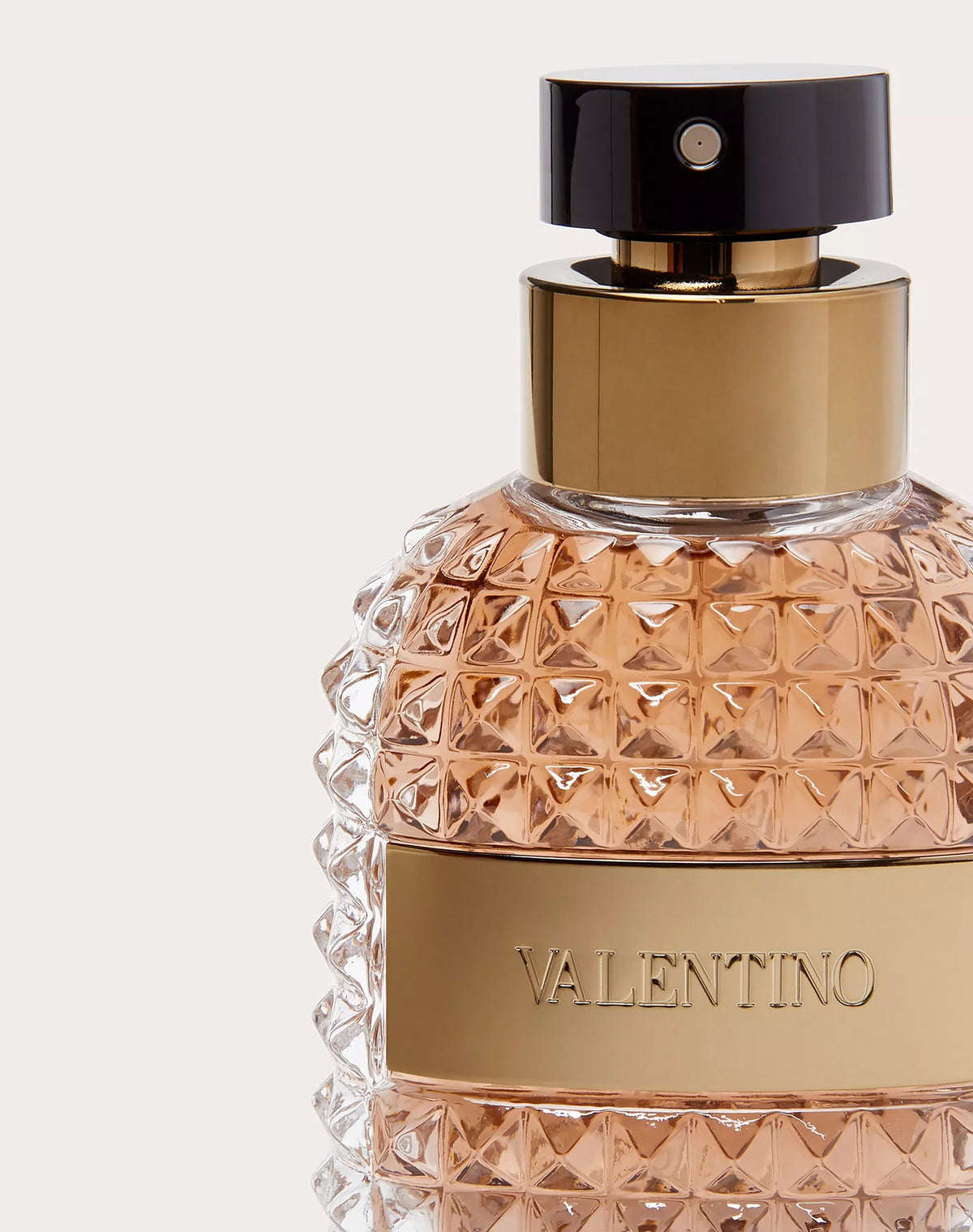 A close-up of a Valentino perfume bottle with a faceted clear glass body filled with peachy rose gold liquid, a gold metallic band with the brand name, a gold neck collar, and a glossy black cap.