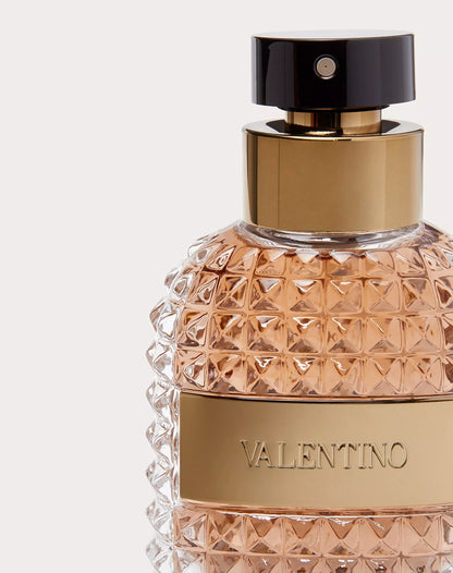 A close-up of a Valentino perfume bottle with a faceted clear glass body filled with peachy rose gold liquid, a gold metallic band with the brand name, a gold neck collar, and a glossy black cap.