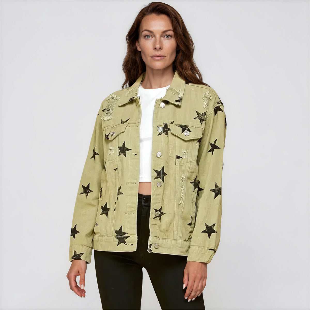 A woman modeling a light olive green denim jacket with black star patches and silver button details, worn over a white crop top and black pants, against a light gray studio background.