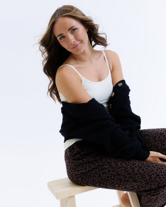 Woman wearing a black cardigan over a white tank top and leopard print pants on a white background