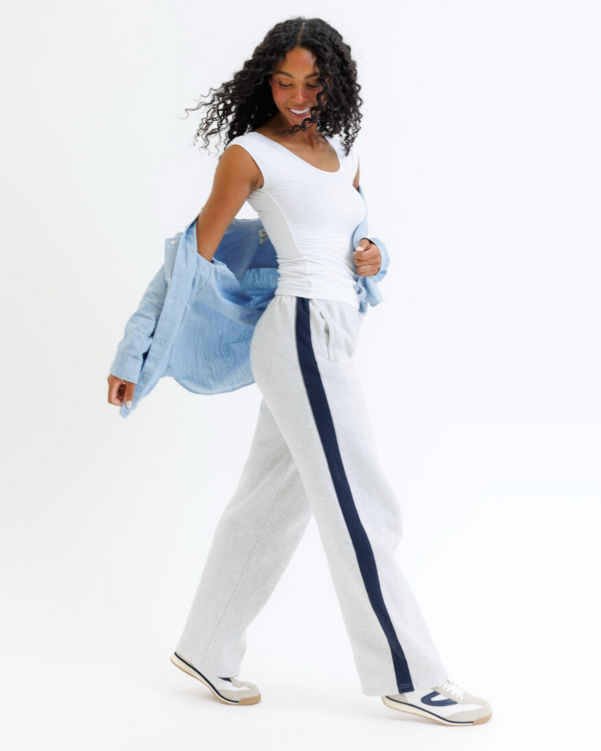 Woman wearing a white top and white pants with a blue stripe, holding a denim jacket.