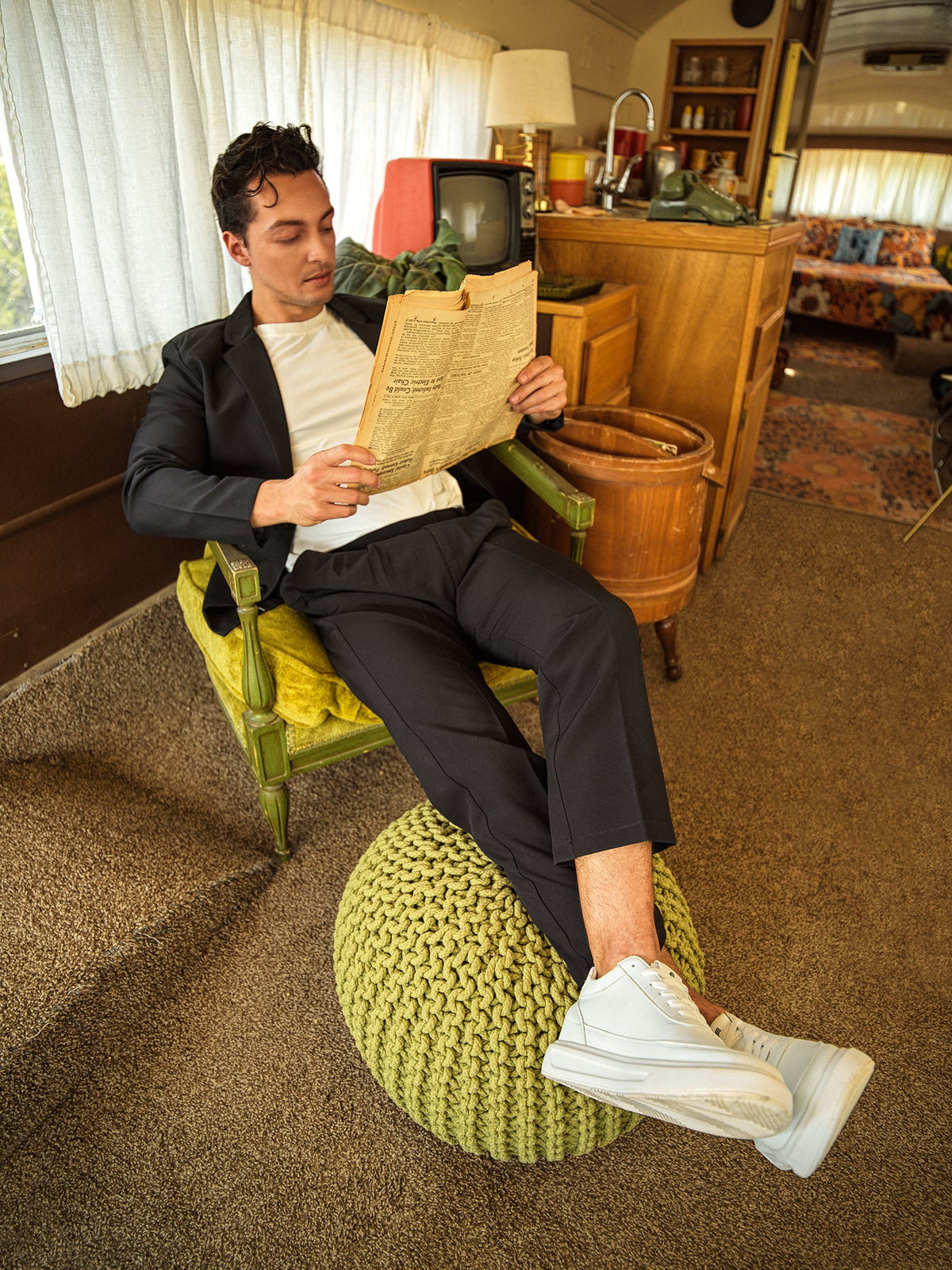 Man sitting on a green ottoman reading a newspaper in a retro-style room.