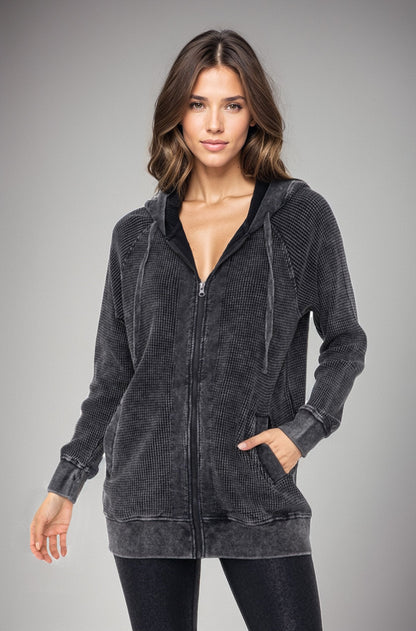 A woman modeling a dark gray washed zip-up hoodie with a textured corduroy-like pattern, black hood lining, and drawstrings, set against a solid gray studio background.