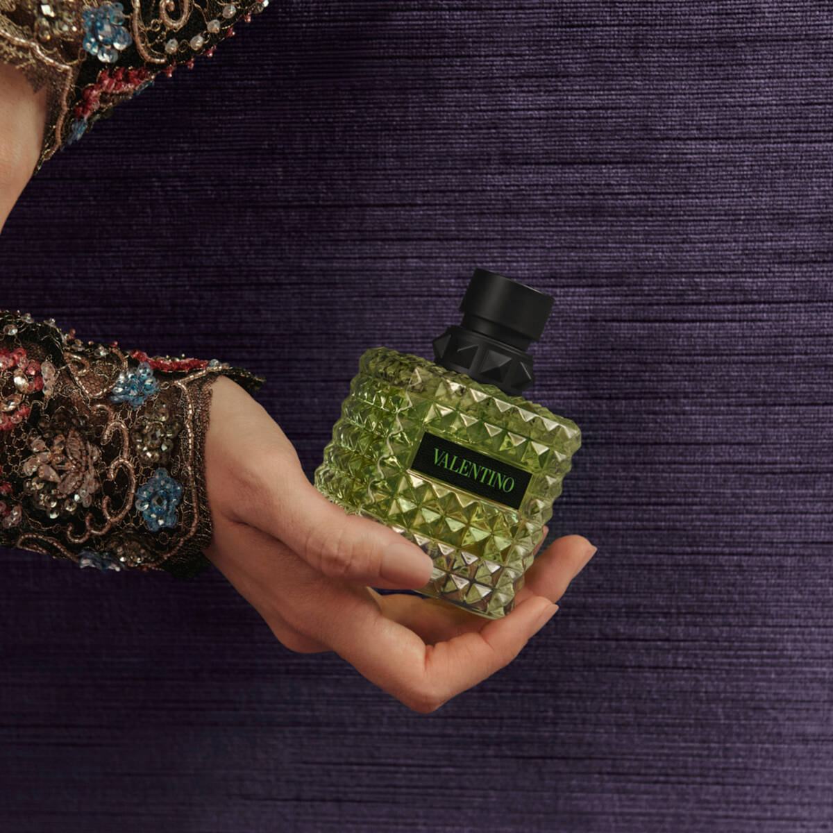 Hand holding a green Valentino Born in Roma Green Stravaganza perfume bottle against a dark textured background.