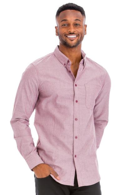 A smiling man wearing a light pink long-sleeve button-down shirt with a chest pocket, paired with black pants, against a solid white background.