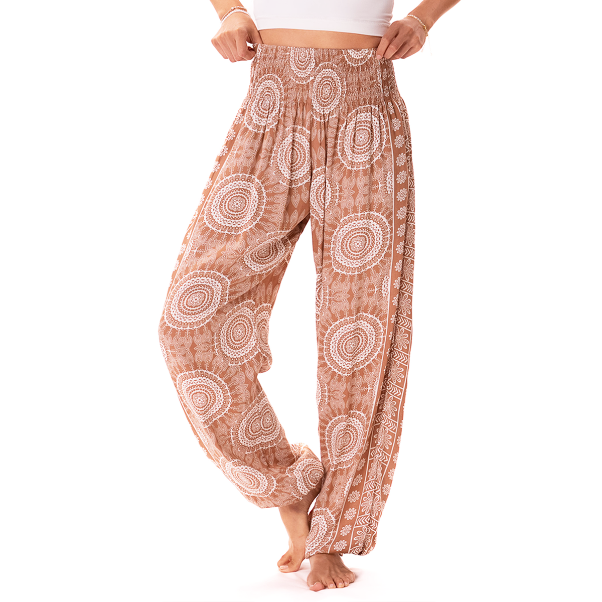 A person modeling loose, high-waisted harem-style pants with an allover white circular mandala pattern on a warm tan background, shown against a plain white studio background.
