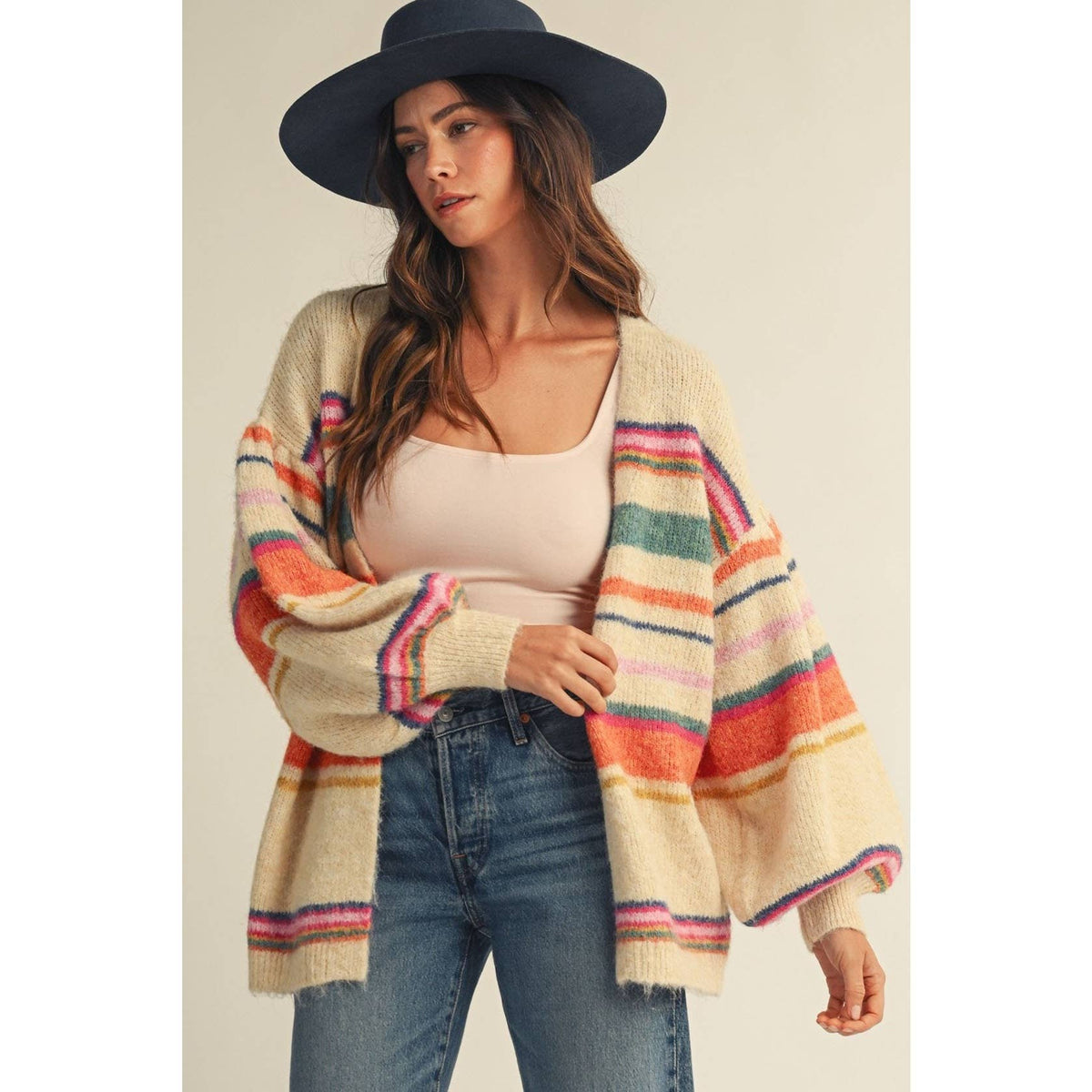 A woman wearing a cream knit cardigan with colorful horizontal stripes in pink, orange, green, and blue, paired with a beige tank top, blue jeans, and a dark navy wide-brim hat, set against a plain beige studio background.