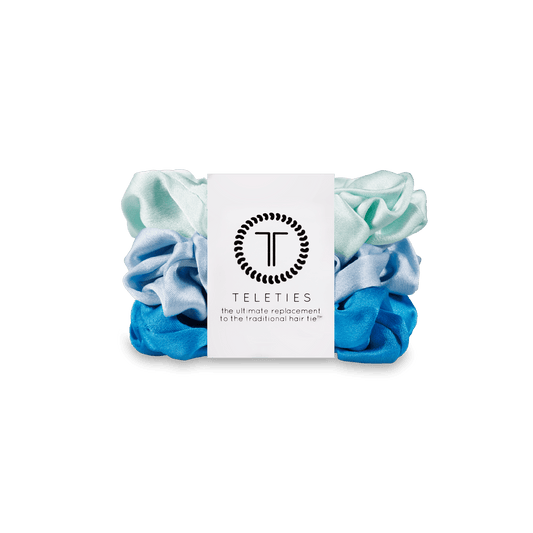 A set of four Teleties hair scrunchies in a blue and mint green gradient, wrapped with a white paper band featuring the black Teleties logo and brand name, set against a solid dark green background.