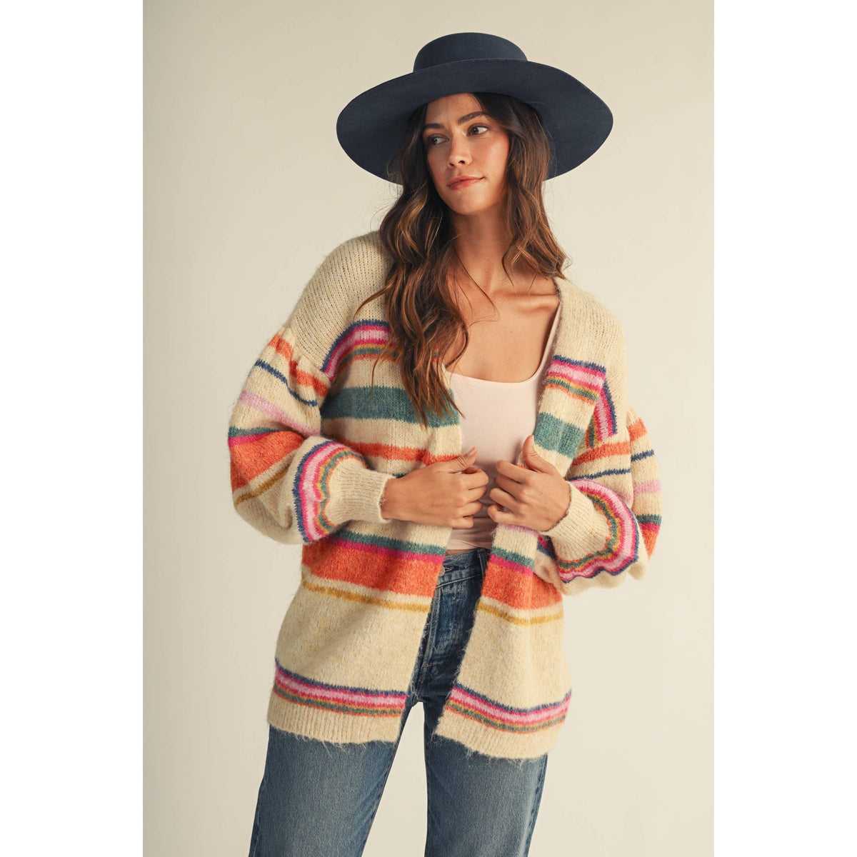 A woman wearing a cream-colored fuzzy open-front cardigan with colorful horizontal stripes in pink, orange, green, and blue, paired with blue jeans and a wide-brimmed dark navy hat, set against a plain beige background.