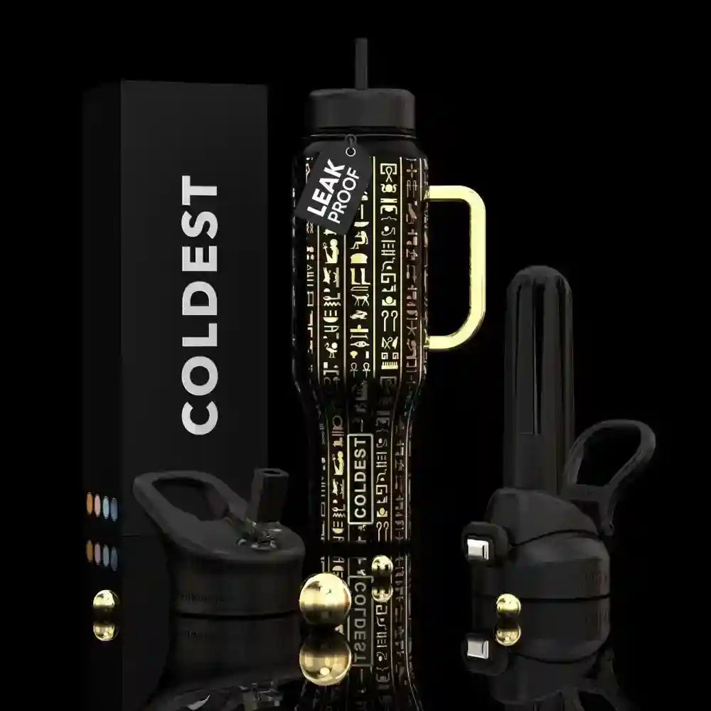 A black insulated tumbler with a gold handle and gold-toned hardware, shown alongside its black branded box and two additional tumbler lids on a black background. The tumbler has vertical gold text reading 'COLDEST' and 'LEAK PROOF'.