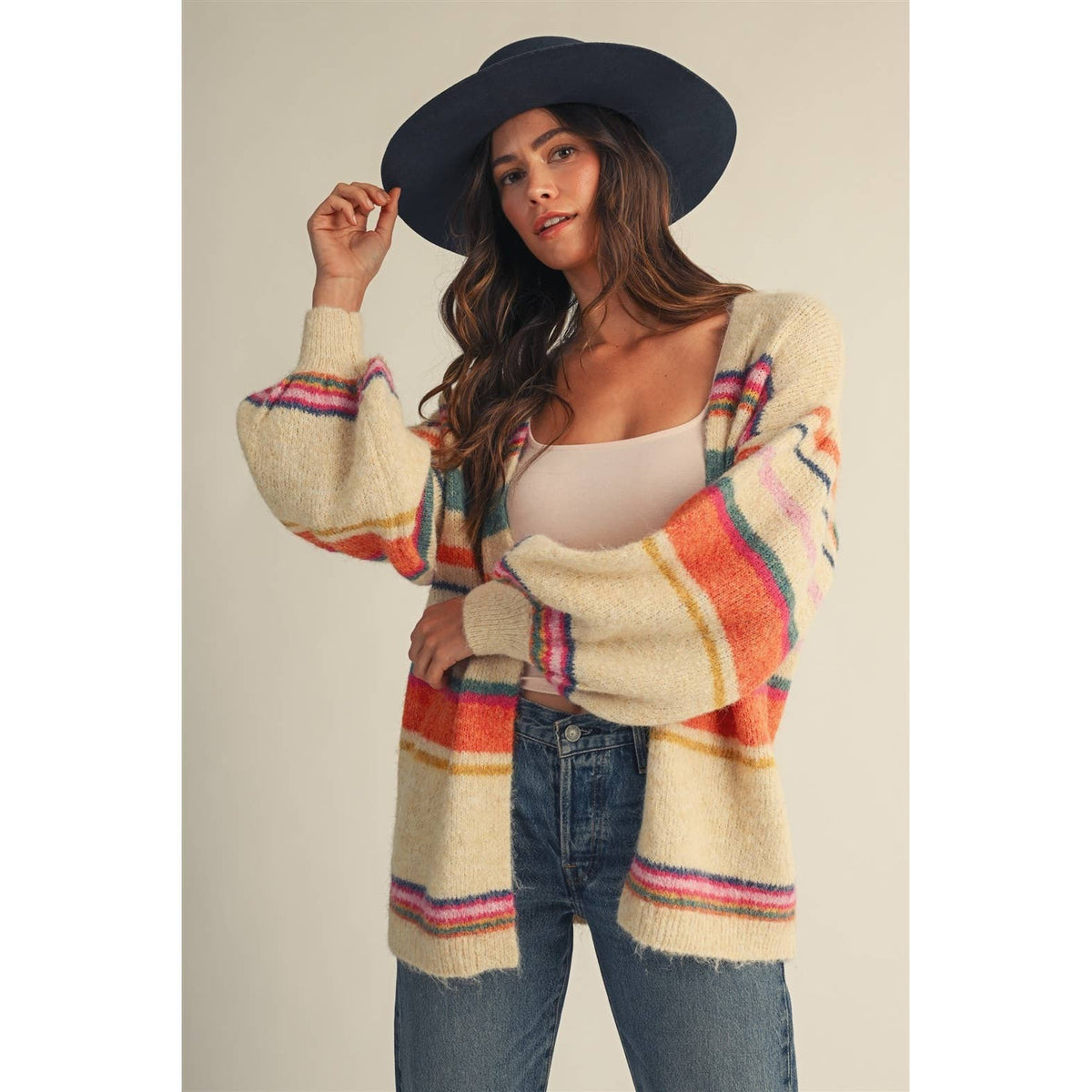A woman wearing a cream-colored fuzzy striped cardigan with pink, orange, and blue stripes, a black wide-brim hat, and blue jeans, posing against a neutral beige background.
