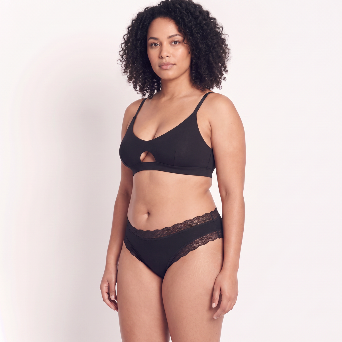 A woman modeling a matching black lingerie set consisting of a bralette with a keyhole cutout and high-waisted briefs with a lace waistband, against a plain light background.