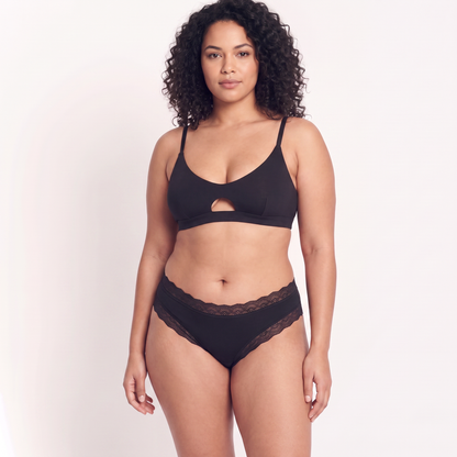 A woman modeling a black two-piece lingerie set consisting of a bralette with a center cutout and matching lace-trimmed panties, against a plain light background.