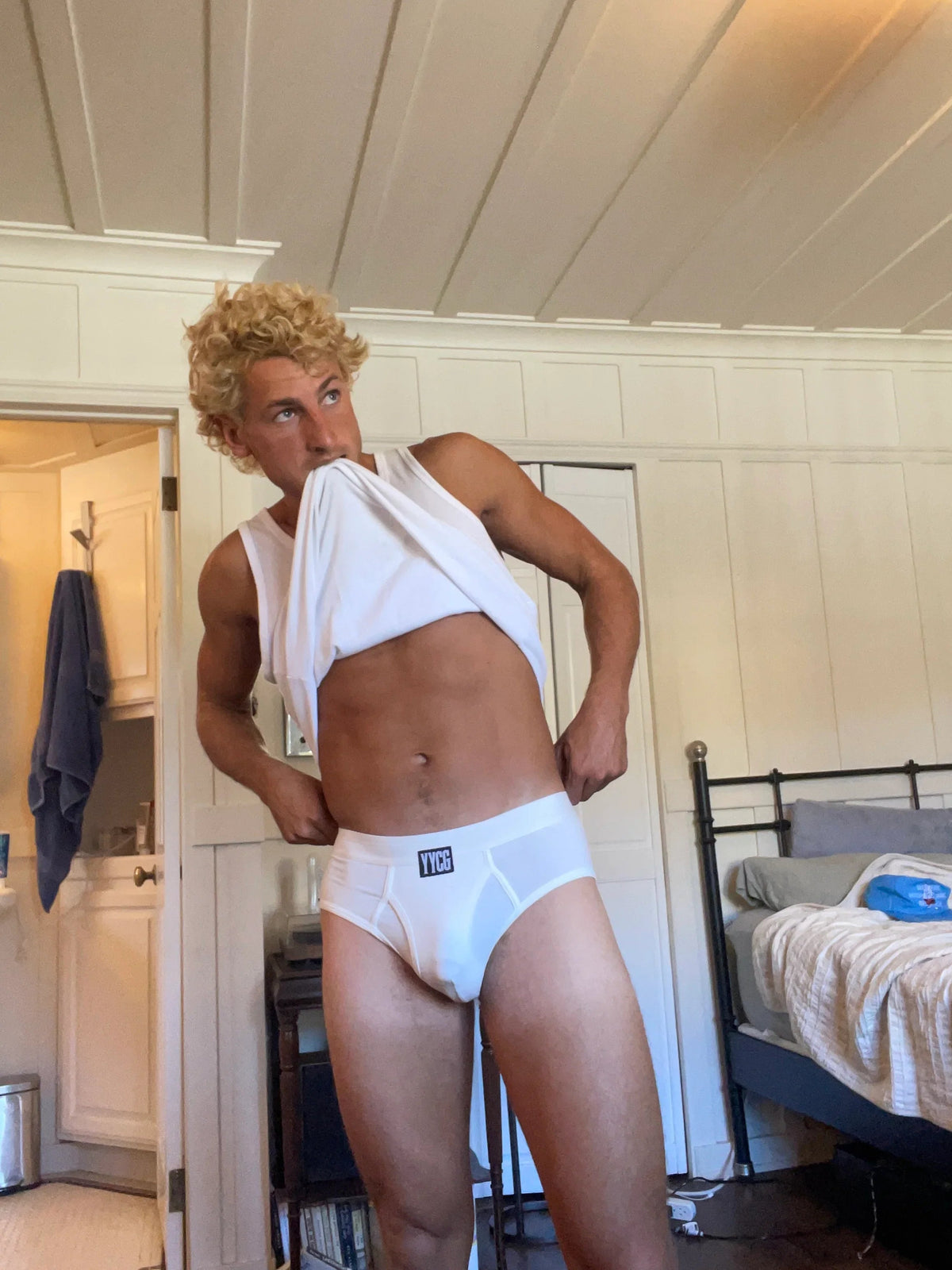 Person wearing a white tank top and underwear in a room with a bed and kitchen area.