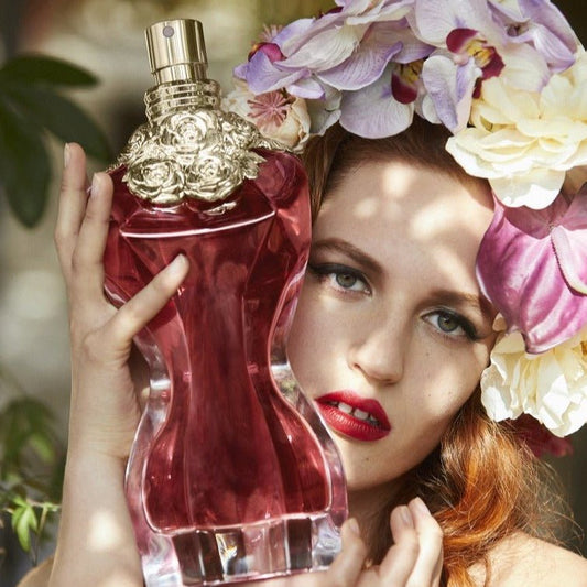 A close-up lifestyle shot of a woman holding a large, sculptural red perfume bottle with a gold cap. She has red lipstick on, and a large floral headpiece of purple and white orchids frames her face.