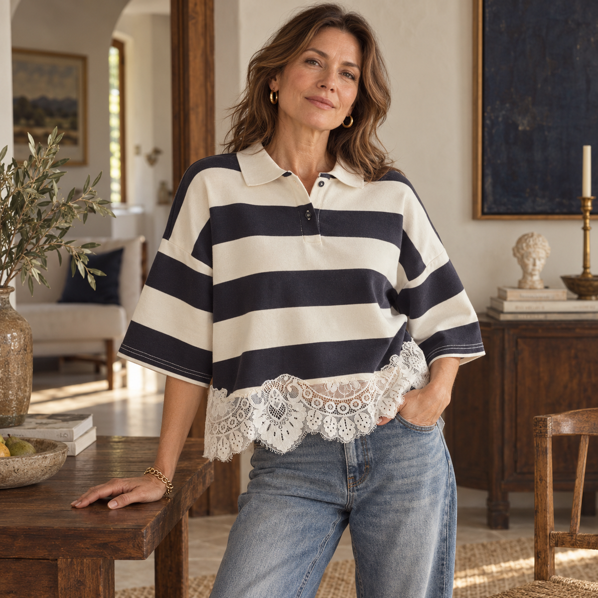 A woman models a short-sleeve polo shirt with bold navy and cream horizontal stripes, featuring a scalloped lace hem and blue jeans, set in a warm, rustic interior room.