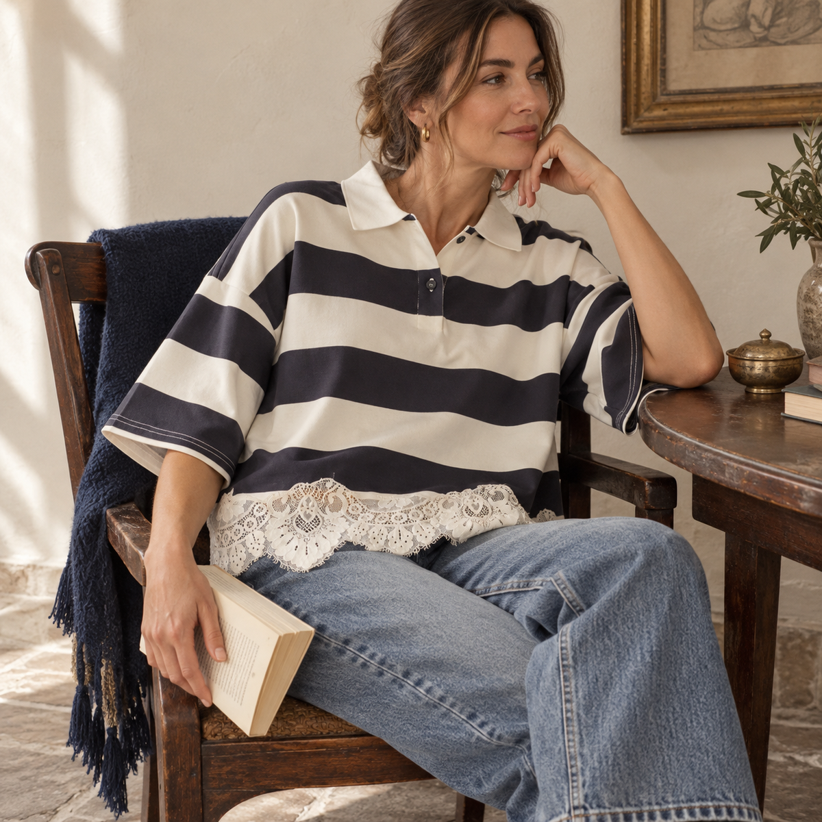 A woman sits on a wooden chair in a sunlit room, wearing a cream and navy striped short-sleeve polo shirt with a lace hem, paired with light blue jeans. A dark blue throw blanket is draped over the chair, and a wooden table with a brass pot is visible beside her.