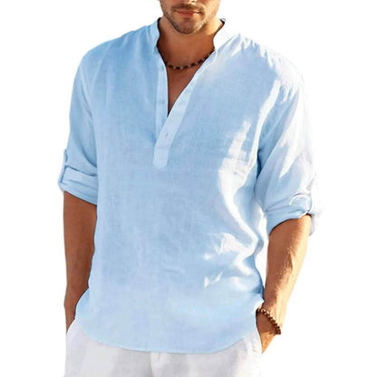 A man wearing a light blue linen henley shirt with rolled-up sleeves and white pants, shown from the chin down against a plain white background.