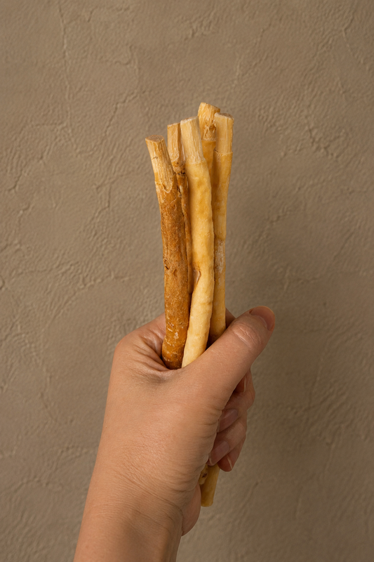 A hand holding a bundle of natural dog chew treats, likely bully sticks or similar rawhide sticks, against a textured beige wall background.