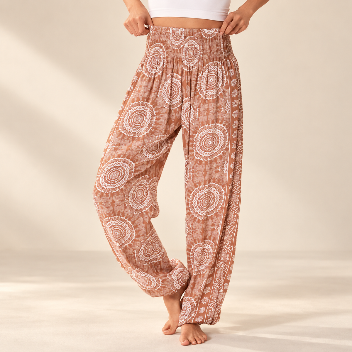 A person wearing loose, high-waisted harem-style pants with a terracotta brown and white circular pattern, standing barefoot against a warm beige studio background.