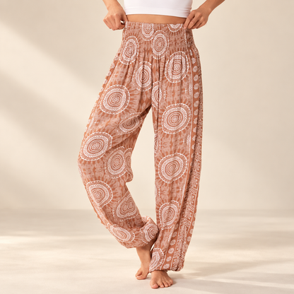 A person wearing loose, high-waisted harem-style pants with a terracotta brown and white circular pattern, standing barefoot against a warm beige studio background.