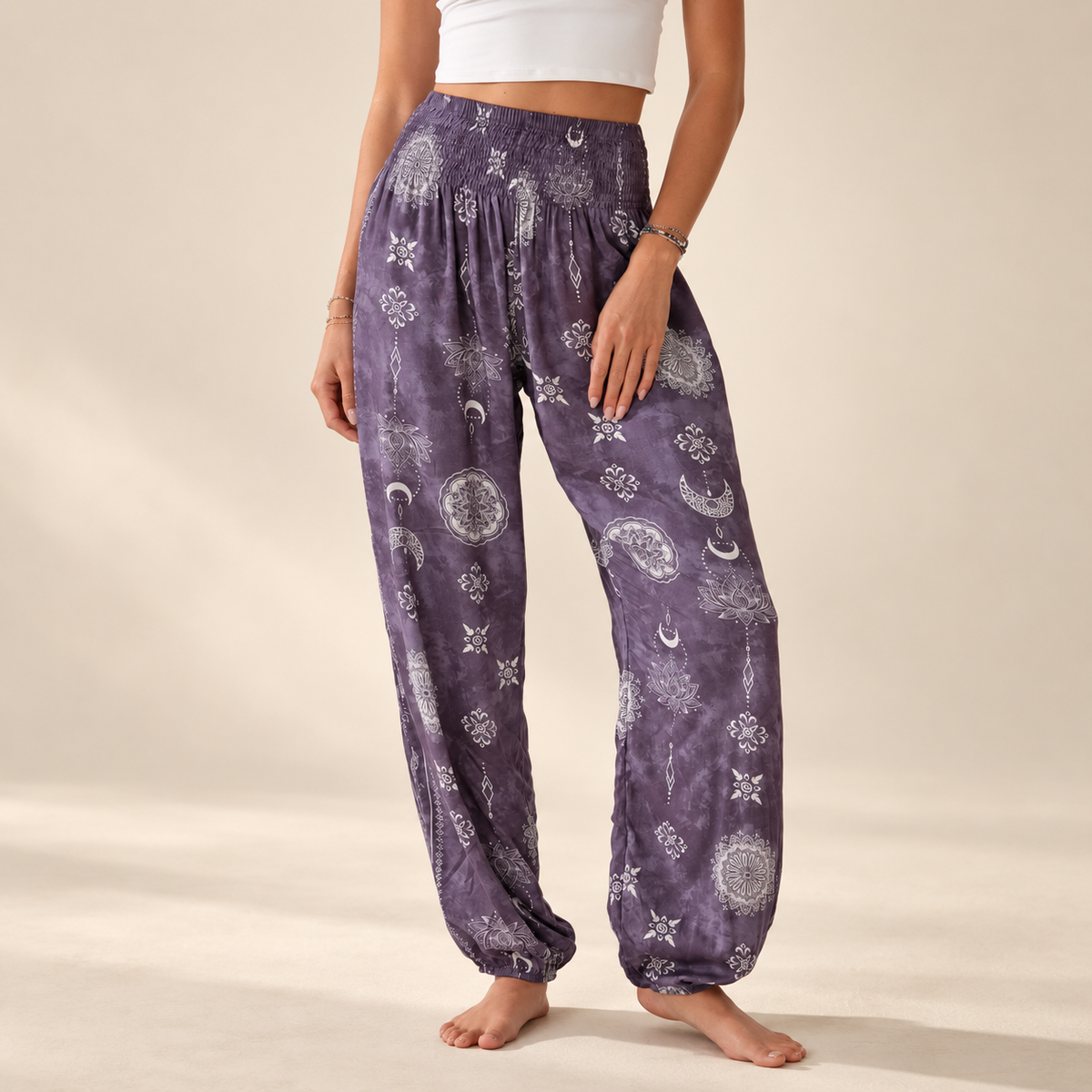 A person wearing loose purple harem-style pants with a white celestial print of moons, stars, and mandalas, standing barefoot against a warm beige studio background.