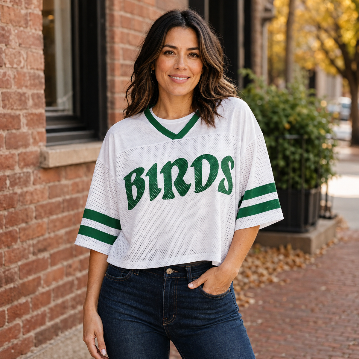 A woman wearing a white mesh sports jersey with green trim and the word 'BIRDS' across the chest, paired with dark blue jeans, standing outdoors in front of a brick building.