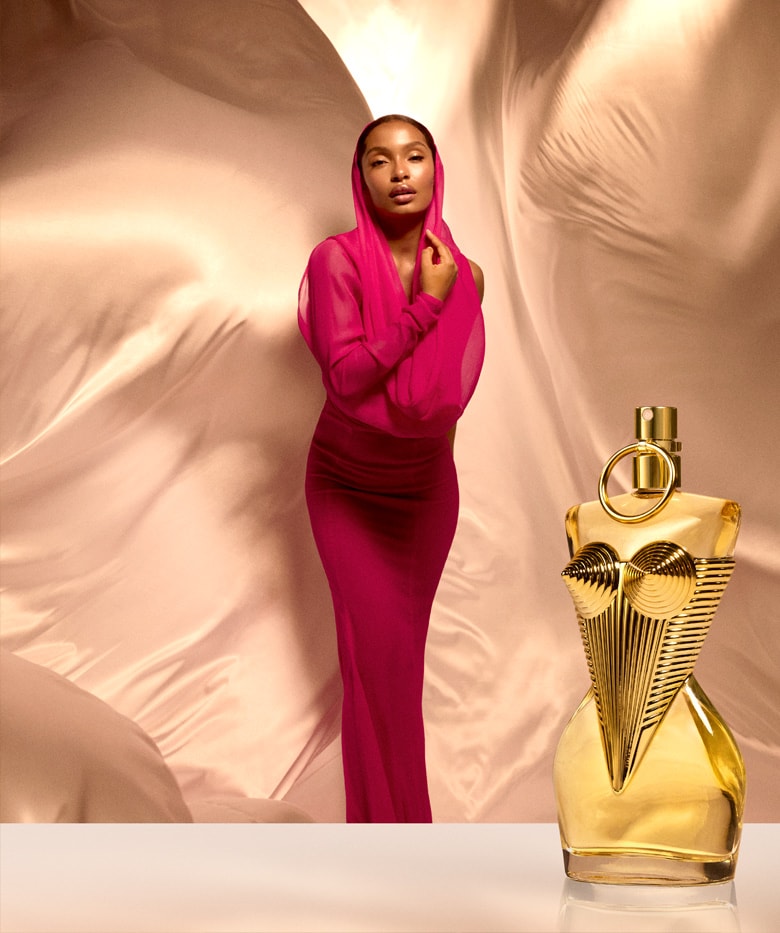 A model in a bright magenta long-sleeve dress stands beside a gold perfume bottle against a draped beige fabric backdrop.