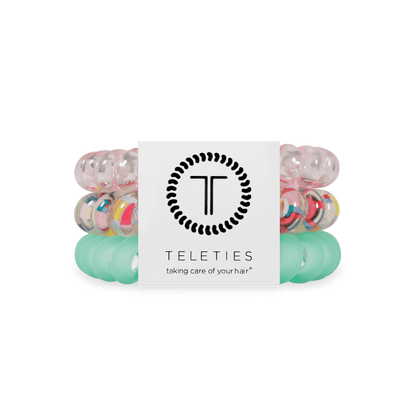 Three Teleties hair elastic bands stacked together with a white paper label in the center. The top band is translucent pink, the middle band is clear with multicolored confetti-like speckles, and the bottom band is translucent mint green.