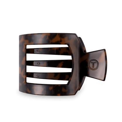A wide, curved hair claw clip in a glossy tortoiseshell pattern, featuring a metal clasp with a circular 'T' logo, set against a solid dark green background.