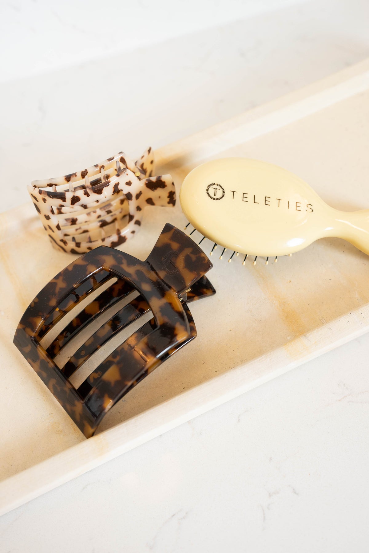 Tortoiseshell hair clip, leopard print bow, and beige brush on a light surface with 'Teleties' branding.
